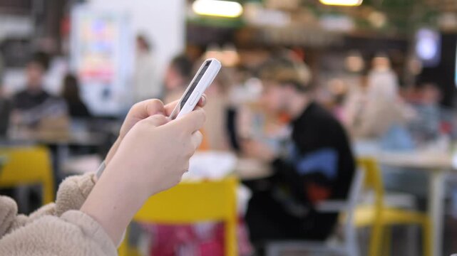Hands hold smartphone in cafe. Blurry background shows people seated. Focused on device, user seems absorbed. Ideal for social media, communication, or technology themes