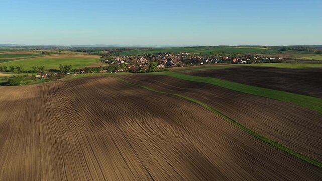 Aerial drone view of a small Czech village nestled among vast rolling plowed fields and green cropland under a clear spring sky, Czech countryside, Moravia, Czech Republic.