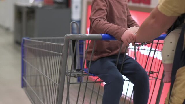 Child sitting in shopping cart holds onto the handle as an adult pushes the cart through a store aisle, with shelves and vibrant displays visible, enhancing the shopping experience
