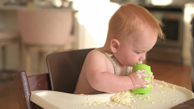 Red-haired baby in high chair engages with a green sippy cup, covered in food remnants, in a bright kitchen filled with soft natural light, creating a warm and inviting atmosphere