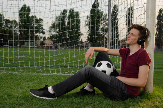 Teenager sitting on the soccer pitch leaned on the goal posing with soccer ball, listening to music