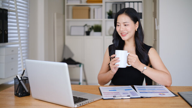 Professional businesswoman enjoying a coffee break while reviewing business charts and documents in a bright office.