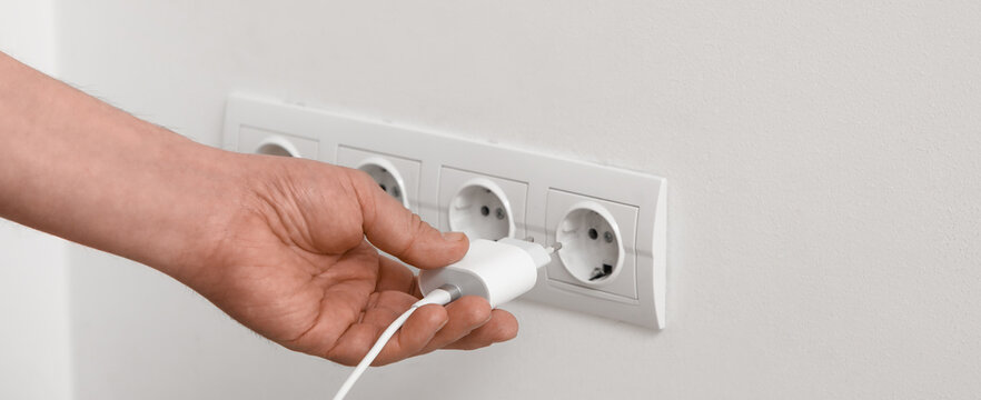 Male hand unplugging charger from socket on light wall at home, closeup