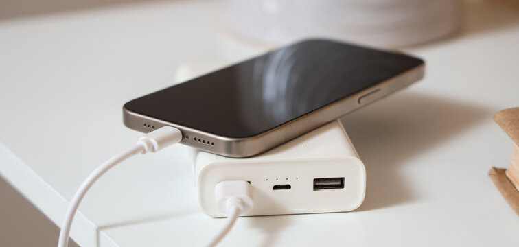 Modern power bank with mobile phone and charging cable on shelf in room