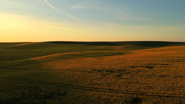 Aerial drone view of gently rolling green and golden farmland at sunset with a wide pale sky and jet contrail above, Moravia, Czech Republic.