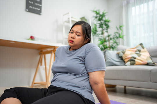 Asian plus-size woman experiencing body aches while exercising in house. 