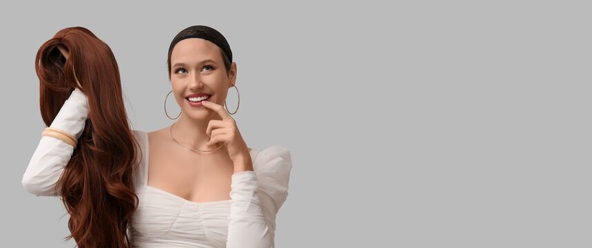 Smiling woman in wig cap holding long brown wig, hair loss and beauty transformation concept, isolated on gray background