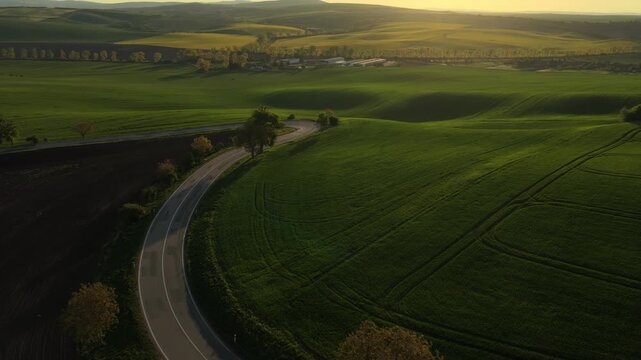 Aerial drone view of rolling green Czech farmland with a winding rural road, farm buildings, trees, and rolling hills at golden hour, Moravia, Czech Republic.