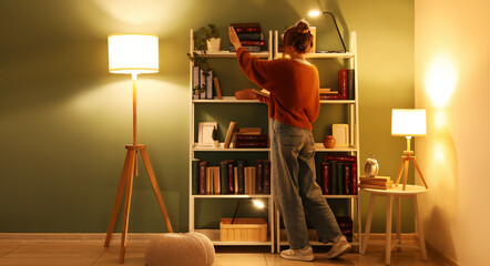Young woman taking book from bookshelves near glowing lamps at home library in evening © Pixel-Shot