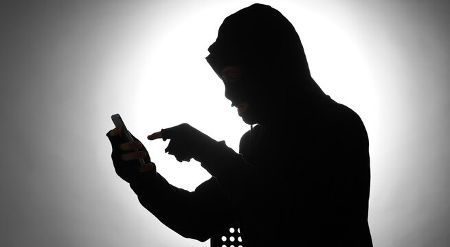 Silhouette of male fraudster with mobile phone at table on light background