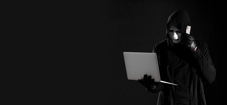 Cybercrime and data theft: anonymous hacker in mask and hoody using laptop and smartphone on dark background, cybercrime and data theft are critical concerns in the digital age