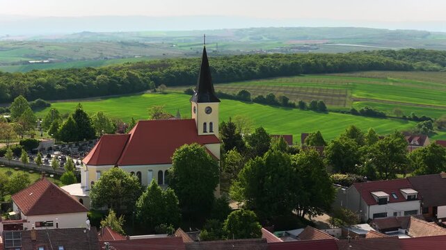 Aerial drone view of a traditional village church with a clock tower and red roof surrounded by trees, with green farmland stretching to rolling hills beyond, Moravia, Czech Republic.