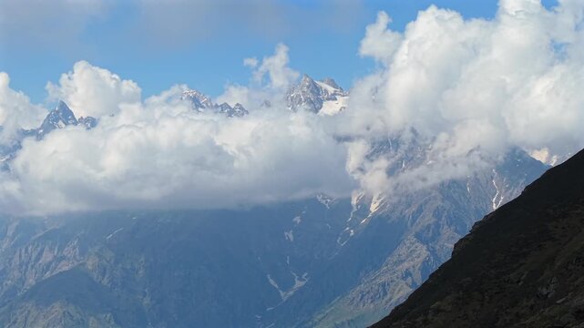 Cinematic aerial of towering snow covered Himalayan mountain peaks emerging through massive rolling clouds beside a rugged rocky ridge near Sar Pass Trek and Parvati Valley, India