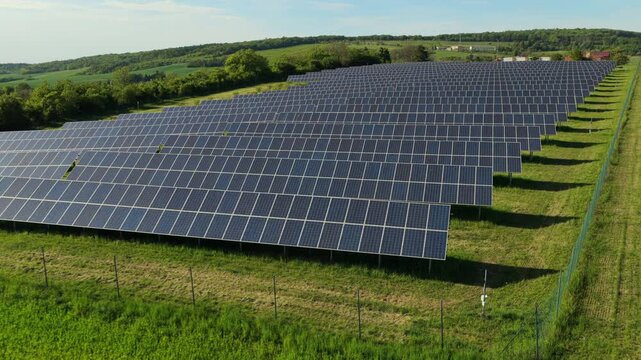 Aerial drone view of a large solar farm with rows of photovoltaic panels set in green grassland beside a forest and rolling hills on a sunny day, Moravia, Czech Republic.
