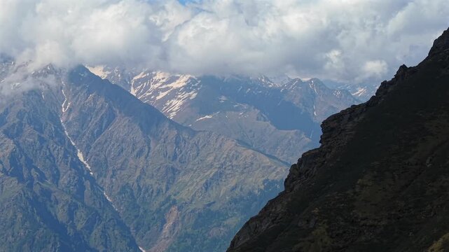Aerial view of dark rocky ridge with snow covered Himalayan mountains and dramatic clouds in the background near Sar Pass Trek and Parvati Valley, India.