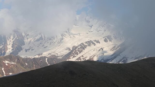 Cinematic aerial view of snow covered Himalayan mountain peaks emerging through dense clouds above rugged alpine ridges near Sar Pass Trek, Himachal Pradesh, Parvati Valley India.