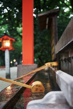 Wooden ladles resting on a stone water basin with a blurry red torii gate and lantern background