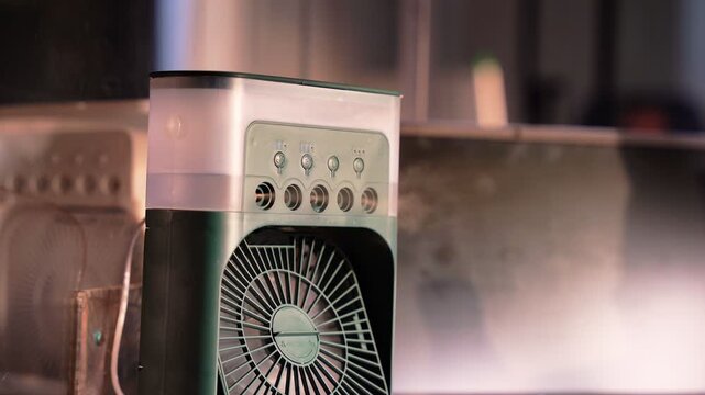 A portable green evaporative air cooler sprays water mist from five nozzles in a dim modern office interior at night near a computer monitor. Cinematic shallow depth of field.