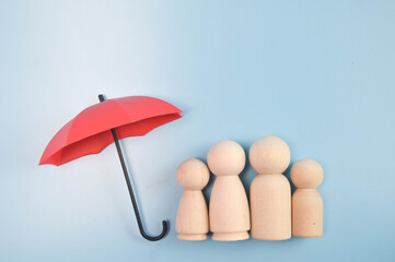 Four wooden peg figures of varying sizes under red umbrella with black handle, symbolizing family protection on light blue background. © YNG