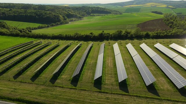 Aerial drone view of a solar farm with gleaming panels amid green countryside and rolling Moravian hills with yellow rapeseed fields in the background, Czech Republic.