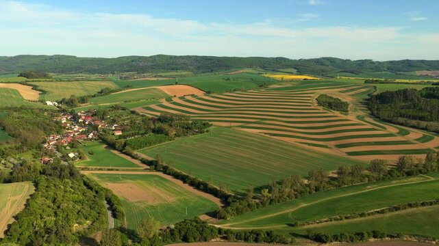 Aerial drone view of rolling Moravian farmland with striking striped crop field patterns, yellow rapeseed patches, a small village and distant forested hills under blue sky, Czech Republic.