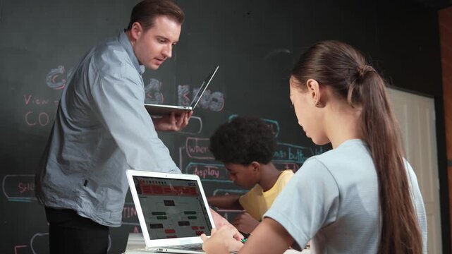 Caucasian girl programming or coding system while african boy fixing model. Teacher holding laptop while looking at diverse children writing web develop program. Innovation education. Edification