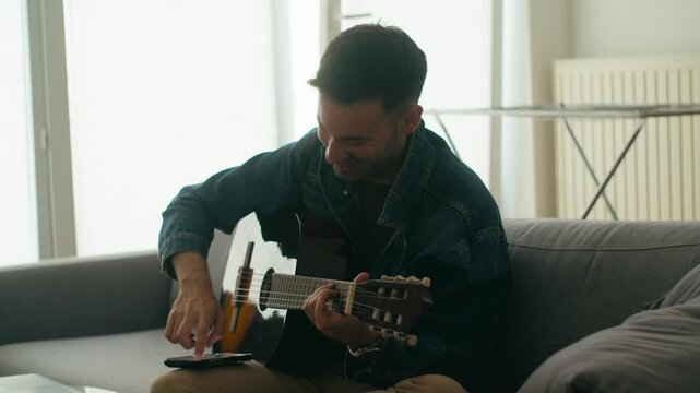 An authentic scene of a young man playing a classic acoustic guitar while sitting on a sofa in a bright apartment. Perfect for lifestyle, music, and wellness content.
