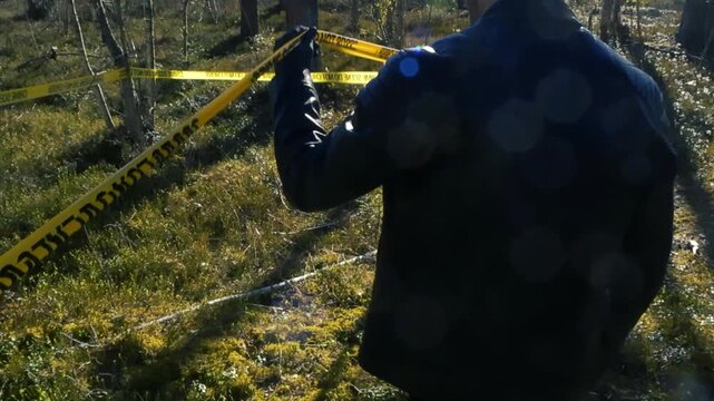 Back view of a police officer in a leather jacket and gloves ducking under a yellow Crime Scene Do Not Cross cordon tape to enter a restricted area of investigation in a sunny forest