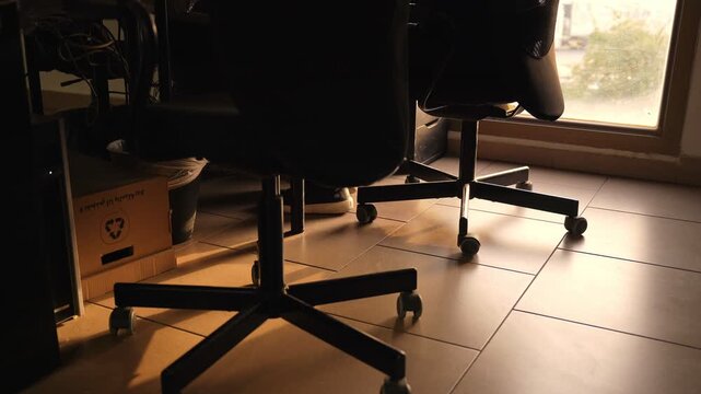 A cinematic low-angle shot of a person sitting in an office chair within a modern office interior. Warm golden hour light highlights the tiled floor, chairs, and a recycling box at dusk.