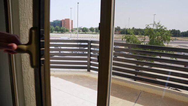 Close up of a man's hand opening and closing a sliding glass balcony door . Overlooking a sunny urban city view with residential buildings and green trees.