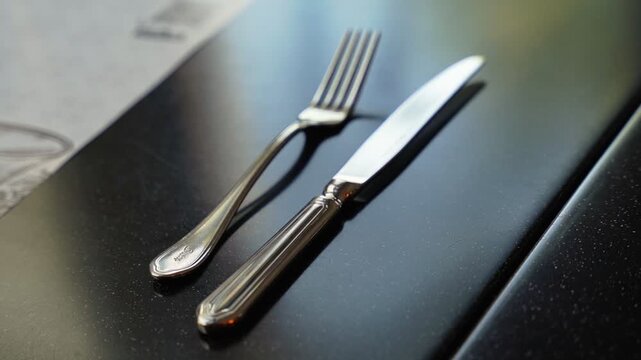 Cinematic close up of a polished silver fork and knife on a dark speckled table during daylight. Blurred restaurant menu background creates a shallow depth of field for dining ads.