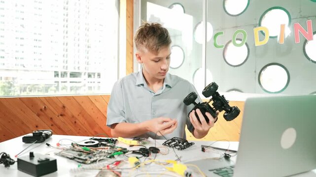 high school boy fixing car model with laptop and electric tool placed on table. Closeup of teenager inspect robotic machine construction while using eletronic equipment in STEM classroom. Edification