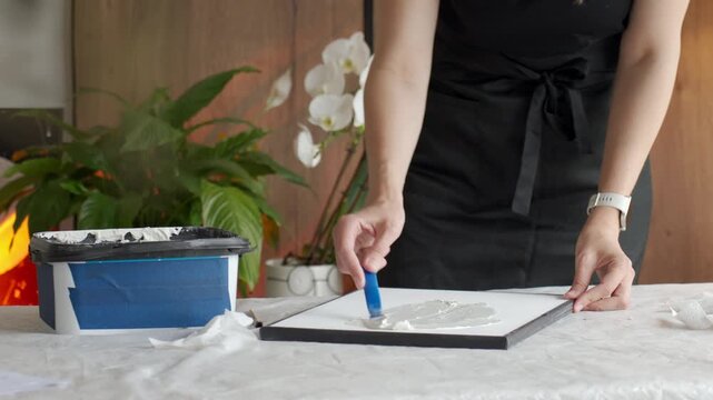 Woman spreads white plaster paste on canvas with a palette knife at a covered work table at home. Concept of DIY textured wall art handmade decor and creative hobby.