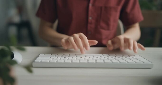 A person wearing red shirt sitting at white desk and typing on computer keyboard at home.