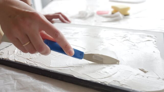 Artist create a textured picture. Hands spreading white decorative plaster with a blue palette knife on canvas in close up. Concept of handmade artwork and creative hobby