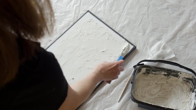 Woman creates a textured picture on canvas applying white plaster paste with a blue palette knife. Concept of handmade artwork and creative hobby.