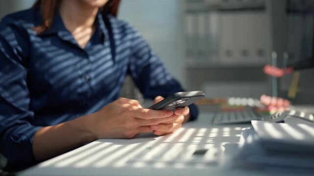 Young woman sitting at a desk using a mobile phone. Striped shadows from the blinds fall on her and the table as she types a message