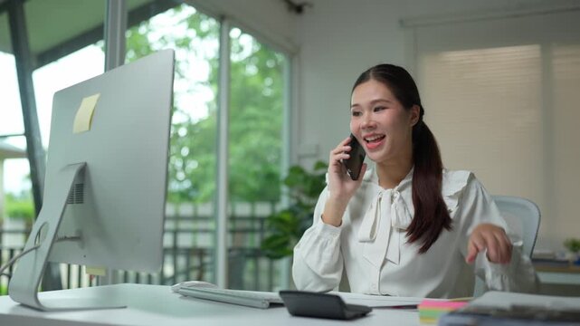 Smiling young businesswoman talking on her cellphone while reviewing documents and using a calculator at her modern office workstation