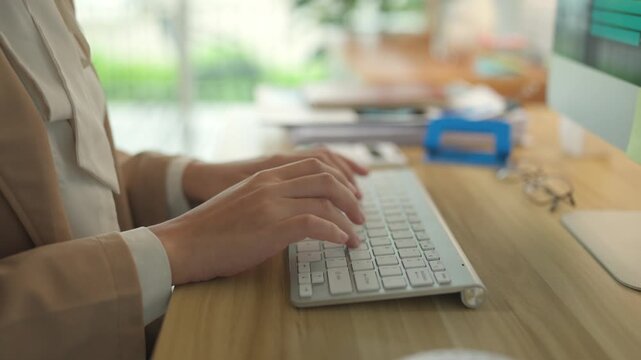 Close up of a businesswoman's hands typing on a wireless computer keyboard. Office worker using a desktop computer at a wooden desk