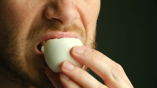 Extreme close up man with reddish beard eating whole hard boiled egg against dark background, showing concept of healthy protein rich food and balanced diet. Bearded man eating a hard boiled egg.
