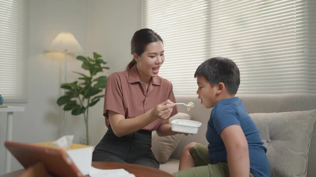 Young caring Asian mother feeding her stubborn little son who refuses to eat. Family lifestyle and motherhood concept in a modern living room