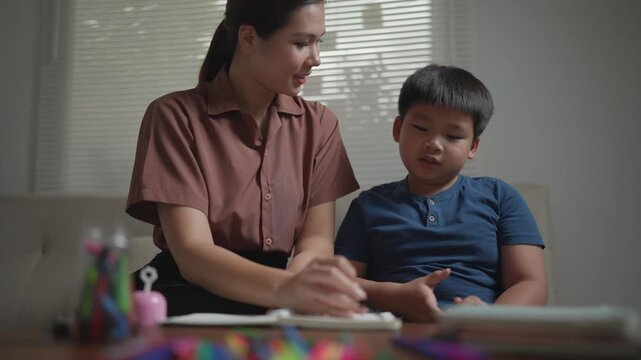 Young asian mother tutoring her elementary school son at home. The boy is learning and writing in his notebook while sitting at the table