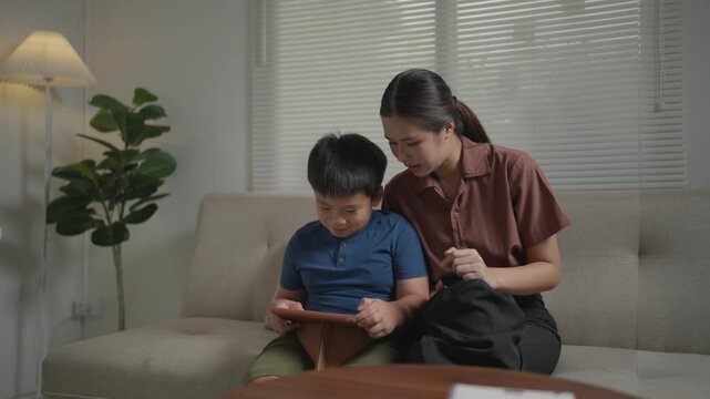 Happy Asian mother and son sitting together on the sofa, smiling and playing educational games on a digital tablet in the living room