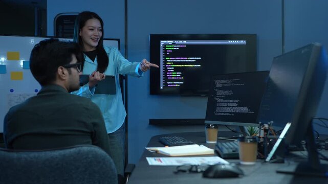 Asian female manager talking to male programmer writing code in dark blue office, Tech team discussing software development, cybersecurity and AI technology, Late night working business team concepts