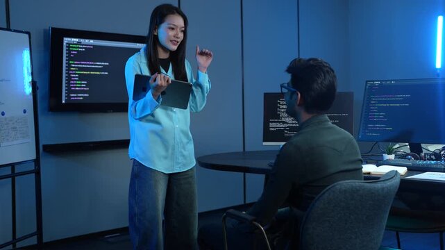 Asian female manager talking to male programmer writing code in dark blue office, Tech team discussing software development, cybersecurity and AI technology, Late night working business team concepts