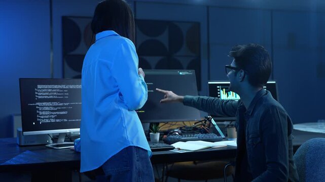 Asian female manager talking to male programmer writing code in dark blue office, Tech team discussing software development, cybersecurity and AI technology, Late night working business team concepts