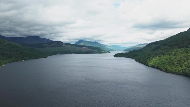 Scenic aerial view of Loch Lomond in the Scottish Highlands. Beautiful drone shot of the vast lake water surrounded by green forest mountains under a cloudy sky.