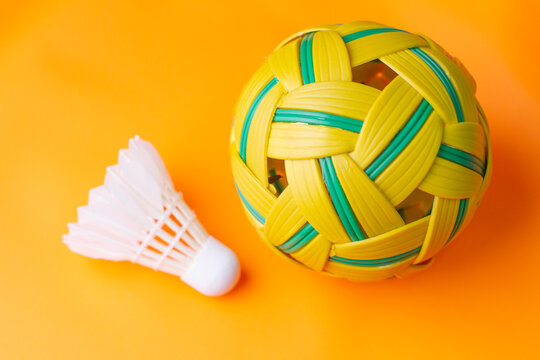 Top view perspective of a white badminton shuttlecock laid horizontally next to a woven sepak takraw ball on a bright orange floor.
