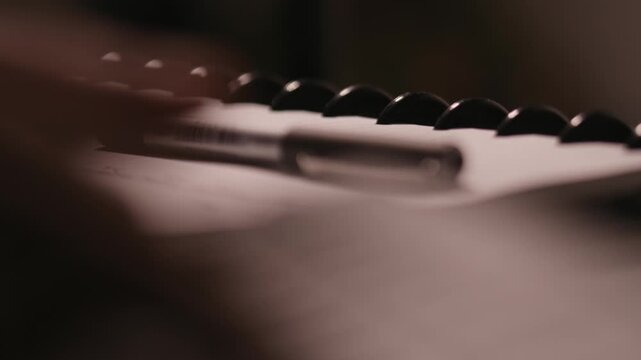 Fingers writing on a keyboard in the blurred foreground with a pen and a notepad in focus in the background.