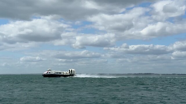 A hovercraft rapidly crosses the Solent waters in England, United Kingdom, under a partly cloudy sky, leaving a spray of water behind it